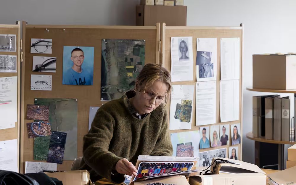 Female investigator in glasses and green sweater reviewing case files at a cluttered desk in front of a large corkboard covered with crime scene photos, suspect and victim images, aerial maps, and police reports in a detective office.