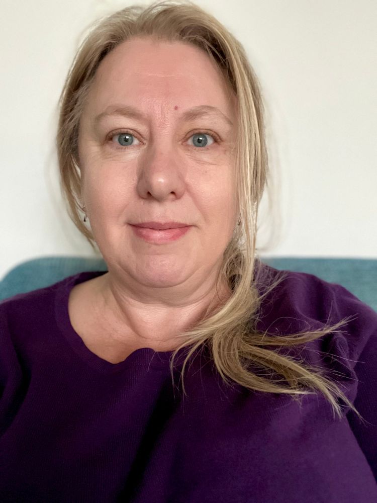 Ik in een paars truitje voor een lichte muur, je ziet nog net een blauwe bankleuning. Ik ben een blonde vrouw van middelbare leeftijd (LOL) met blauwe ogen - mijn haar springt uit een losse vlecht! 