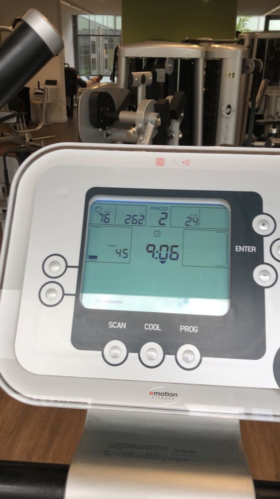 A close-up view of the screen and controls of an Emotion Fitness exercise machine, likely a cycle, with the display showing numerical values for time, distance, and other workout metrics. The background is blurred but shows other gym equipment.
