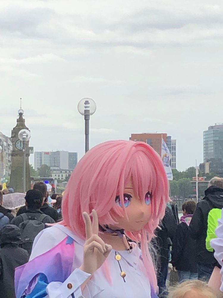 Eine Person mit einer pinken Perücke und einer großen Anime-Maske mit weiten lila Augen hält zwei Finger zum Friedenszeichen hoch. Sie trägt ein weißes Oberteil mit farbigen Schulterakzenten. Im Hintergrund sind eine Menschenmenge, einige Gebäude und ein Uhrturm zu sehen. Der Himmel ist bedeckt.
