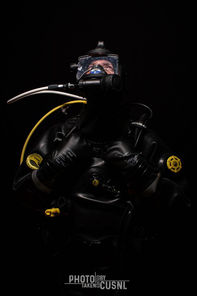 A man looks up into the bright light while wearing a black rubber Viking drysuit. His rubber glove covered hands are grabbing his dive vest. Contrastful yellow elements of his diving equipment show in the picture.