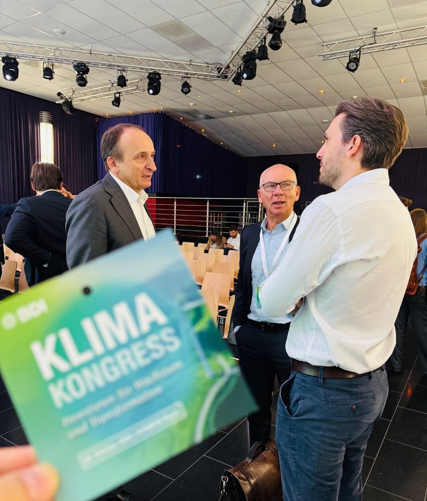 Drei Personen im Gespräch beim BDI-Klimakongress, im Vordergrund ein Teilnehmer mit Kongress-Badge „Investitionen für Wachstum und Dekarbonisierung“; im Hintergrund Stuhlreihen und Bühnenbereich.