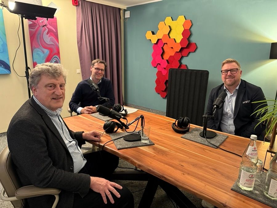 Drei Personen sitzen an einem rechteckigen Holztisch in einem modernen Podcast- oder Interview-Setup. Auf dem Tisch stehen mehrere Mikrofone, Kopfhörer und Glasuntersetzer. Im Hintergrund sind farbenfrohe Wanddekorationen zu sehen, darunter eine Kunstinstallation aus roten und gelben hexagonalen Elementen sowie abstrakte Gemälde. Eine Wasserflasche und ein Glas stehen im Vordergrund auf dem Tisch. Die Szene ist professionell ausgeleuchtet, mit einem Studiolicht auf der linken Seite.