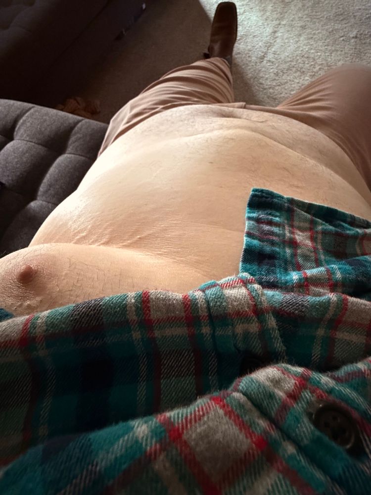 Larger man sitting back with shirt lifted to expose belly and one nipple. 