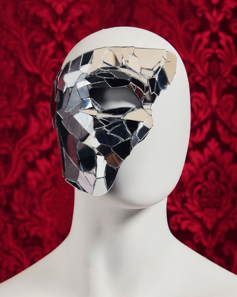 Phantom of the Opera half mask which covers the right side of the face, constructed from shattered mirror pieces and displayed on a white mannequin head. The background is an ornate red floral wallpaper. Photo from Vogue Magazine.