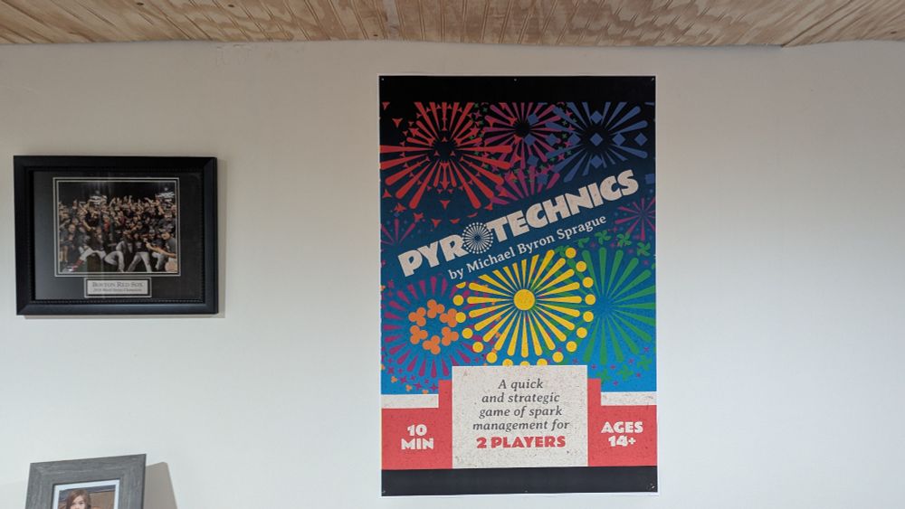 Poster of the cover for upcoming game, Pyrotechnics hung in the office basement of the designer. Art shows fireworks in the games art style and the title is tilted left across the middle. 