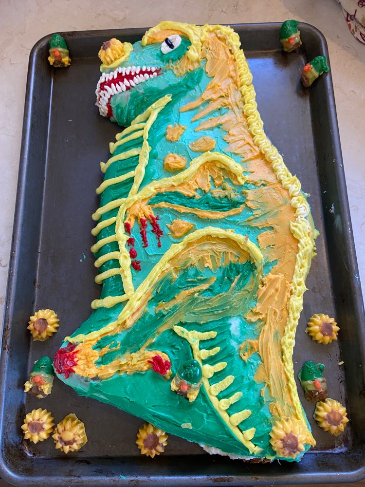 Dinosaur sheet cake poorly decorated, with candy gnomes and flowers around it.