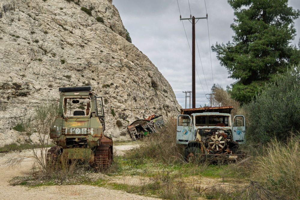 Abandoned quarry with rockface on the left with trees and foliage on the right. The main subjects are an abandoned bulldozer on the left and an abandoned truck with its doors open on the right. In the middle distance is a derelict wagon. Electricity cables and pole lead off into the distance.