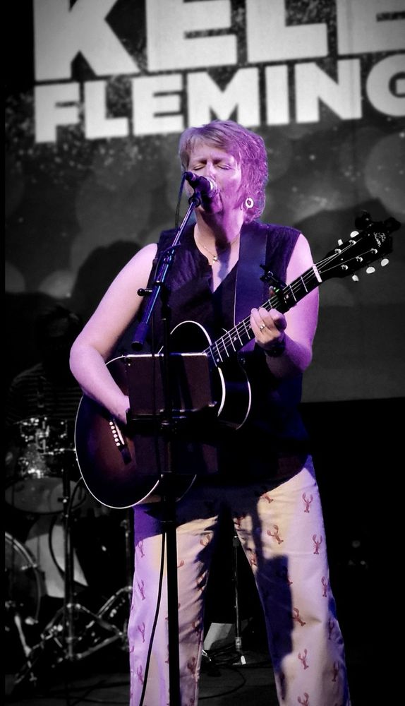 A photo of a woman with red hair singing and playing guitar onstage. She’s wearing a black tank top and pants with a lobster print. 