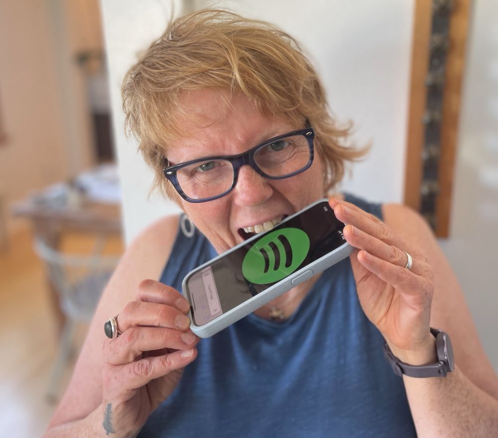 A photo of me - a white woman with short red hair and freckles wearing a blue tank and blue glasses - taking a bite out of an iPhone displaying the Spotify logo. 