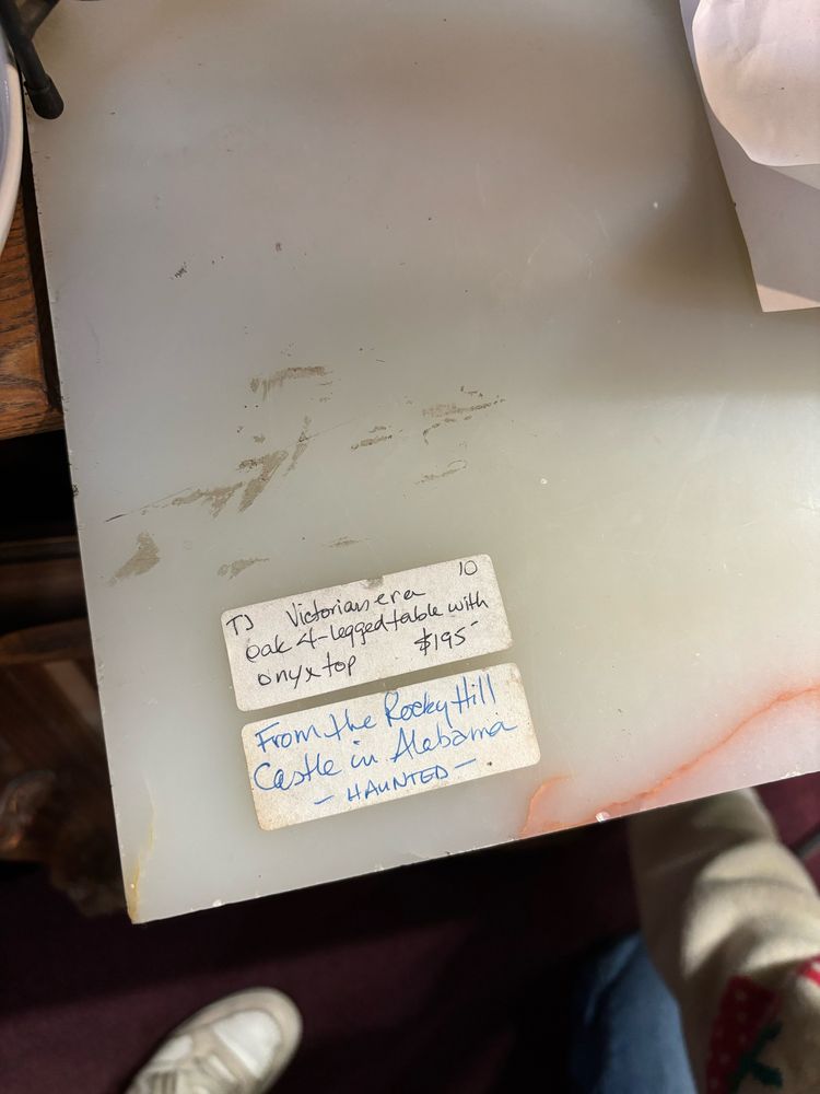 a table in an antique shop with two stickers that say “victorian era oak 4-legged table with onyx top $195” and “from the rocky hill castle in alabama - HAUNTED”