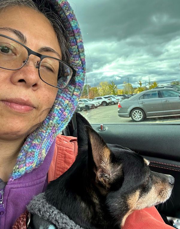 Half-blind brown woman (me) in glasses, holding Mi Madre’s Chihuahua/miniature Doberman pincher dog while seated inside a car. Parked cars, trees in fall foliage, and grey cloudy sky visible in background. 

Really appreciate how Mi Madre added that crocheted-trim to the edge of my hoodie after the damn thing shrank in the wash; now it warmly covers my head again.