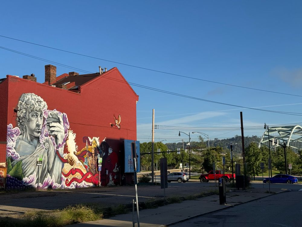 Giant mural painting of a Greek statue-like man on a solid red brick colored building against a bright blue morning sky, low verdant green hills in background and the scaffolding of Pittsburgh’s Hot Metal Bridge across the Monongahela river at right side. Some car traffic (perpendicular to viewer) also visible as are some electric service wires