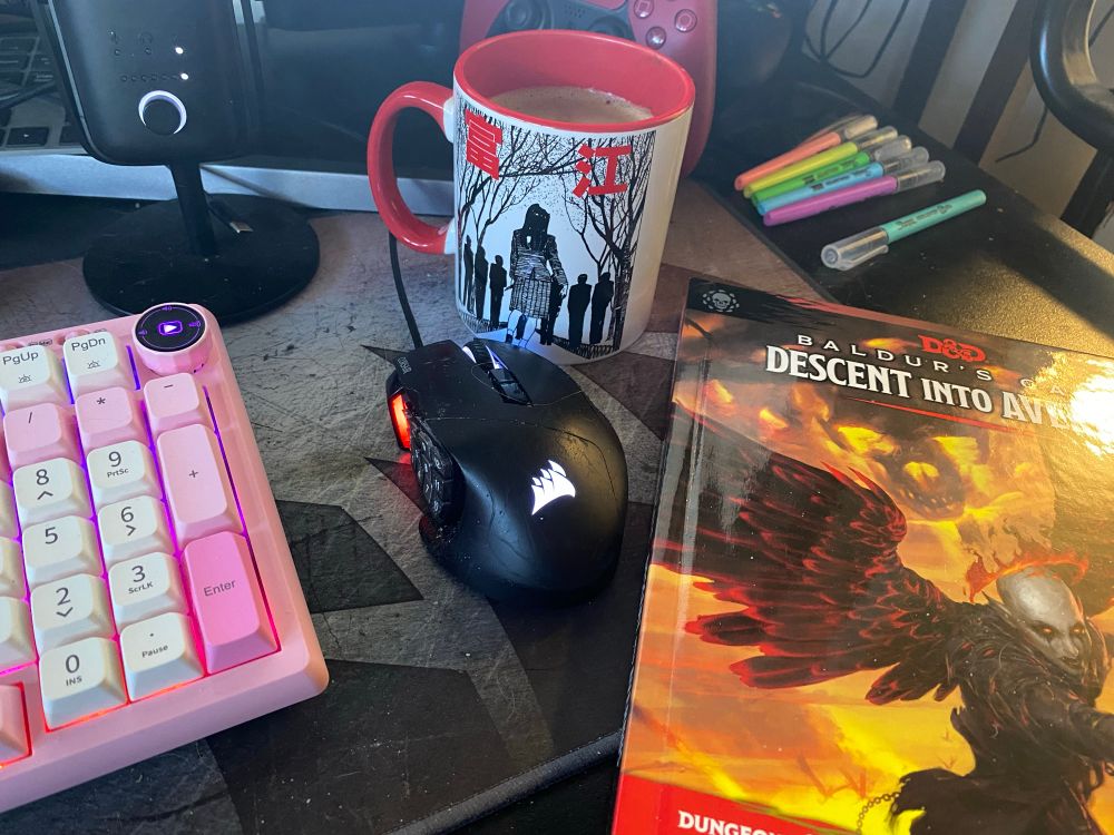 An image of a computer desk with various items on it including a keyboard, mouse, microphone, coffee mug, and Descent into Avernus Dungeons and Dragons campaign book.