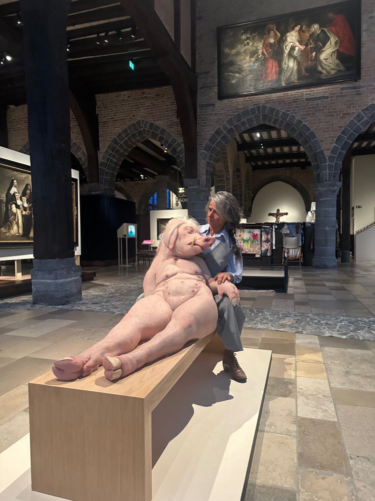 A hyper realistic sculpture. A modern nun with long grey hair sits aside a bench. A pig-human figure, clearly unwell, lies on the bench and leans into her lap. The nun looks at her with kindness and concern. This sculpture is in the middle of a museum in an old religious hospital