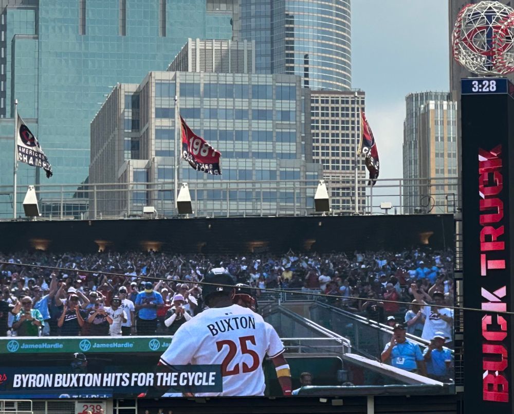 zoomed-in shot of the left field scoreboard at Target Field, with flags on the wall and the city skyline in the background. the board says “BYRON BUXTON HITS FOE THE CYCLE” and shows #25 Buxton from the back