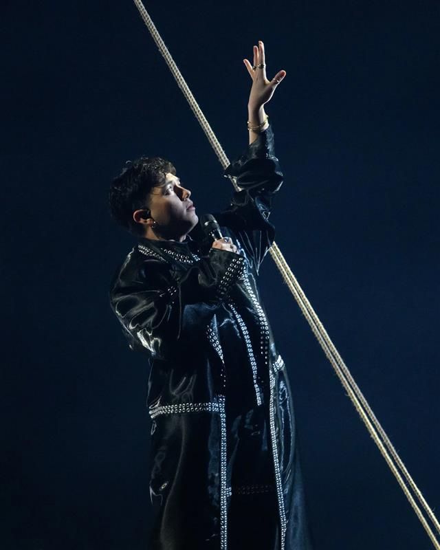 A person on stage wearing a long, dark outfit holds a microphone and raises one hand upward. A thick rope runs diagonally in the background against a dark backdrop.