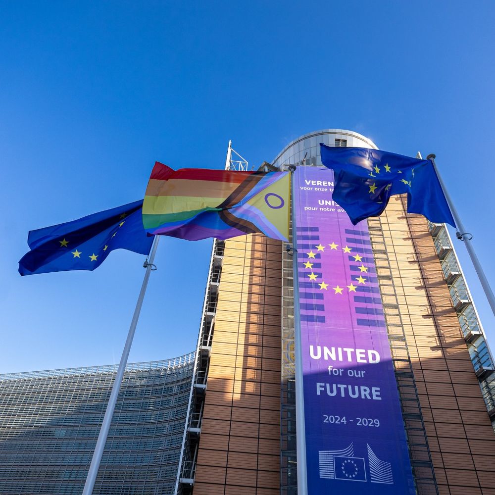 The progressive flag flies between two European Union flags in front of the European Commission headquarters in Brussels. Behind them, a large purple banner on the Berlaymont building reads “United for our Future 2024–2029” with a circle of yellow stars forming the EU symbol.