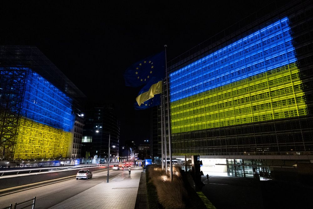 The buildings of the European Commission and the European Council are illuminated in blue and yellow, the colours of the Ukrainian flag. Between them, the street, an EU flag and a Ukrainian flag are visible. It is otherwise dark. 