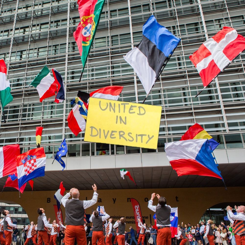 A group of people dressed in coordinated outfits are gathered in front of the Berlaymont building of the European Commission in Brussels. They are tossing various European national flags into the air in celebration. A large yellow sign in the centre reads "UNITED IN DIVERSITY," echoing the European Union's motto. Spectators watch the vibrant and festive scene from the background. 