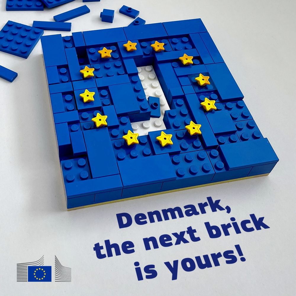 A LEGO brick mosaic depicts a blue rectangle with a white cutout in the shape of Denmark. Yellow star-shaped LEGO bricks are placed on the blue sections, representing the stars of the European Union flag. Below the mosaic, a slogan reads "Denmark, the next brick is yours!" in a blue, handwritten-style font. In the bottom left corner, the logo of the European Commission is visible. 