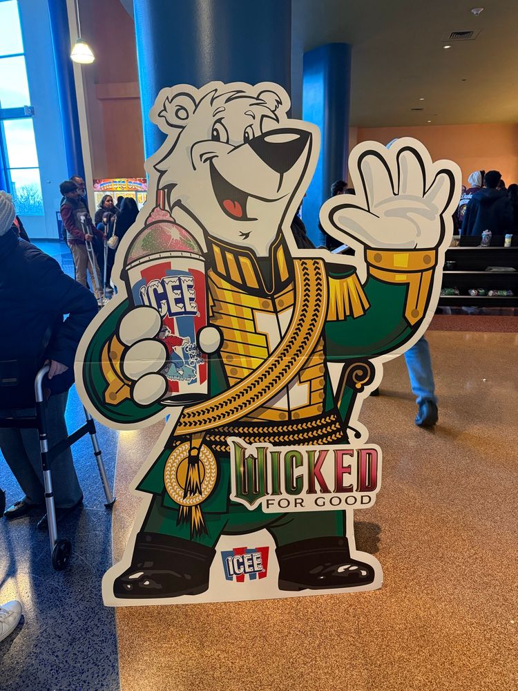 The icee bear, a large cartoon polar bear, dressed in a green and gold coat, waving and holding an icee. Around about his waist is the text Wicked For Good. 