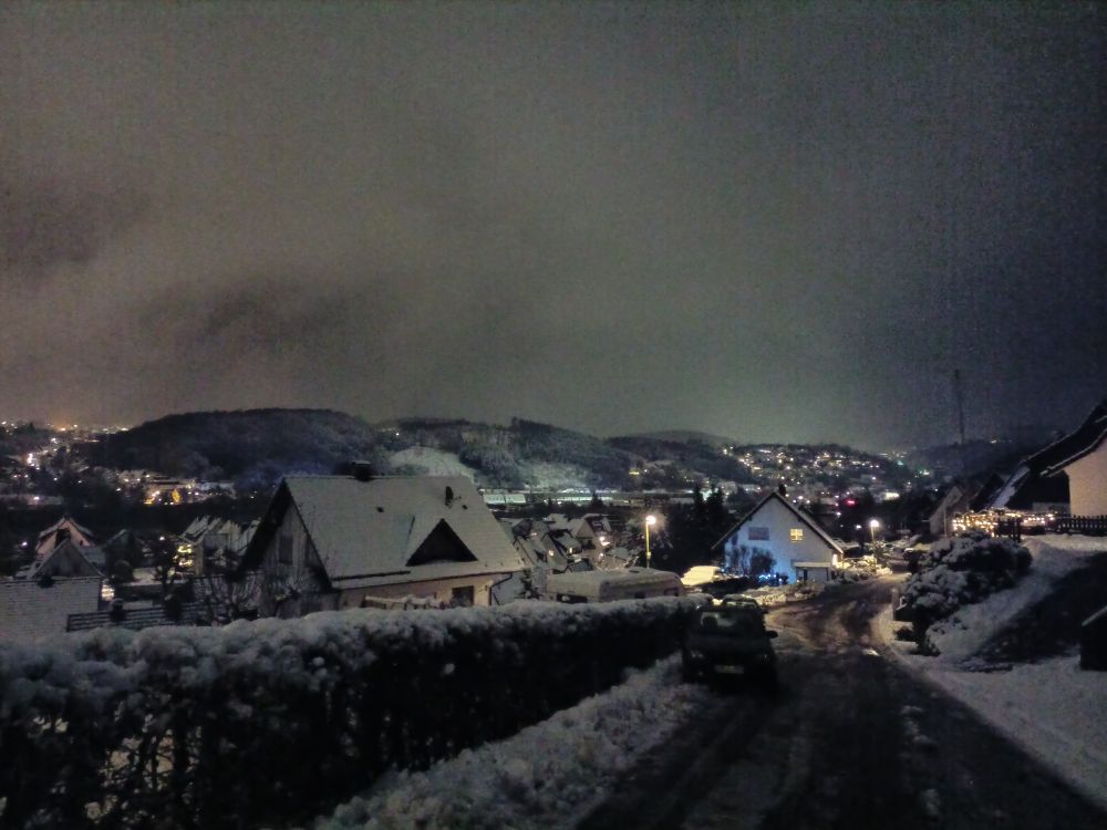 Nachtaufnahme von einem Hügel aus in einem Wohngebiet bei Schnee mit Blick auf die umliegenden Häuser und Berge.