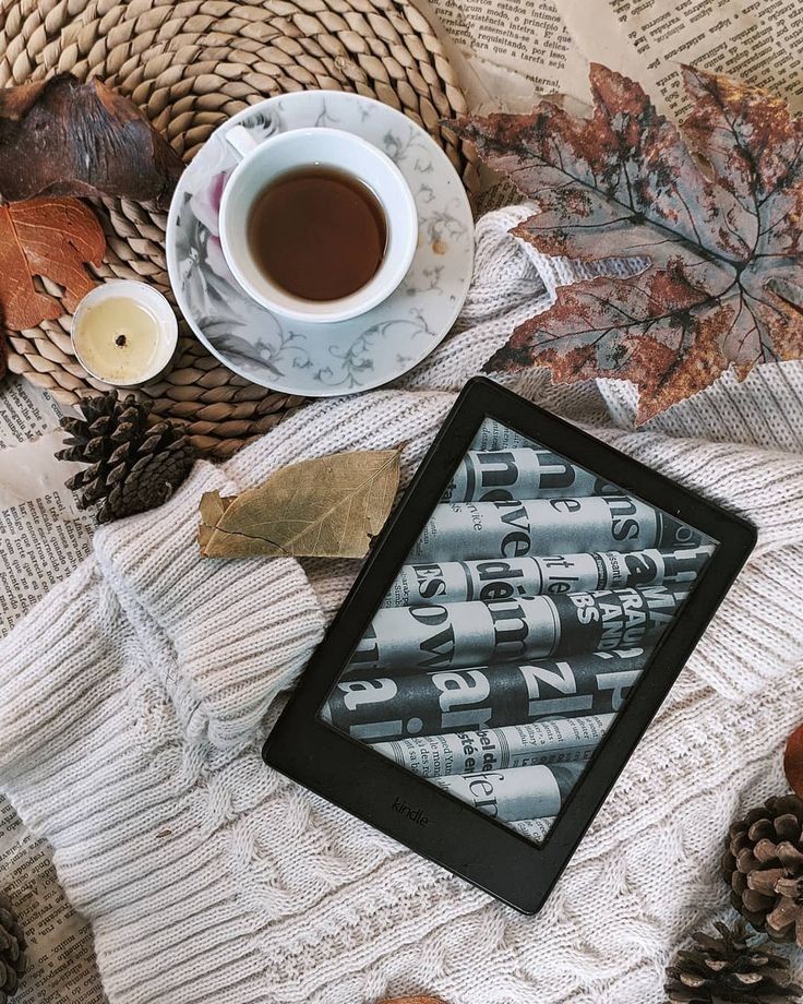 imagem de um kindle em tela de descanso, repousado sobre um suéter bege, com folhas de livros, uma xícara de café e uma vela ao redor 