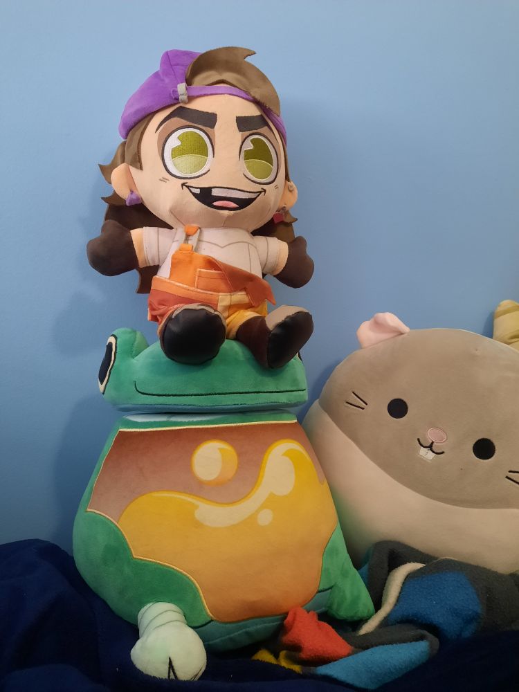 A plushie of Frankie sitting on top of a larger plushie of Boombox, both from the animation 'Knights of Guinevere'. Next to them is an unrelated rat Squishmallow plushie.