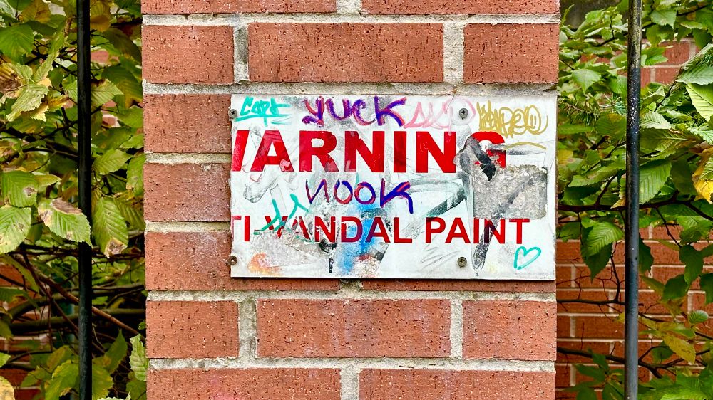 An official-looking council sign with red letters that reads “WARNING ANTI-VANDAL PAINT”, part of which has been broken off, with the remaining part covered in graffiti.