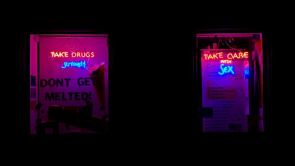 Surrounded by darkness, two shop windows are lit up with neon signs that read “Take drugs seriously” and “Take care with sex”.