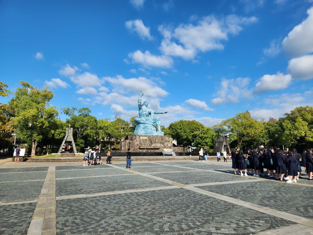 A blue statue of a man, seated, one arm pointing up and another pointing to his left. This is the Nagasaki peace statue that is located in Peace Park