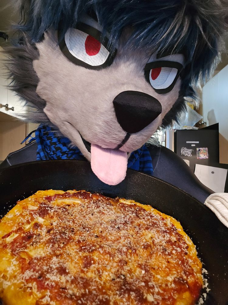 wolf bravely posing with a hot pizza in a cast iron pan, looming over that pizza with a tongue dangling over