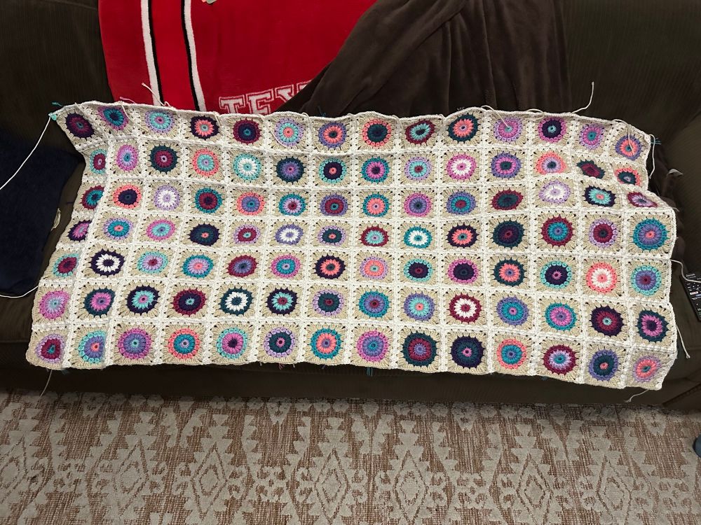 Picture of slightly more than half a blanket made up of individually crocheted starburst granny squares in shades of pink, blue, and purple with white and off-white borders