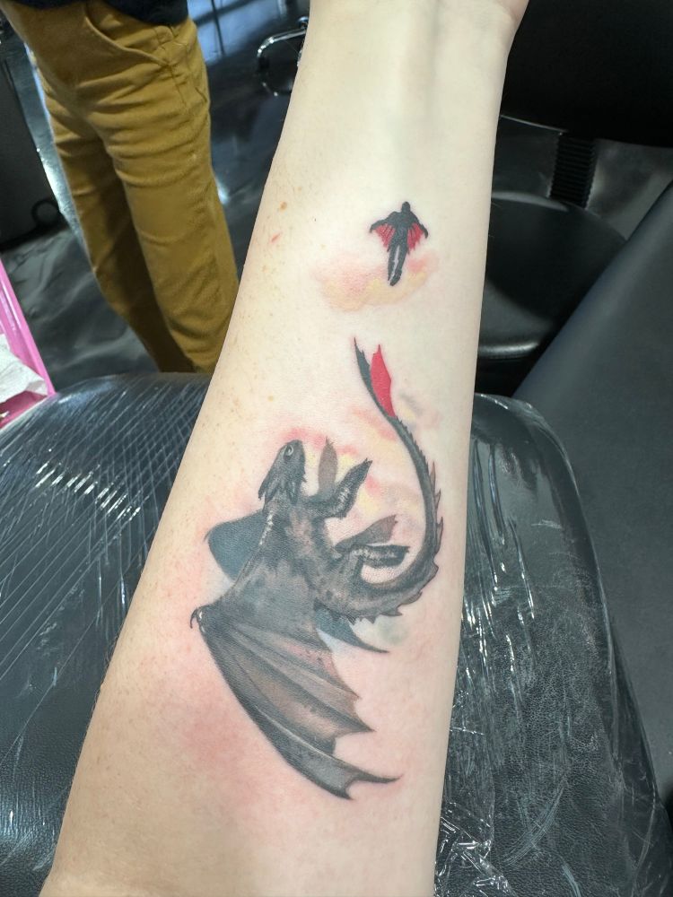 Fresh tattoo on the forearm of a fair-skinned woman, still red from irritation. The tattoo depicts a black dragon with one red tail fin behind a silhouetted figure of a person wearing a red wing suit, with the person’s lower left leg missing. In the backdrop are light watercolors in pinks, blues, and yellows. The characters depicted are Hiccup the person and Toothless the dragon from the How to Train Your Dragon animated movies.