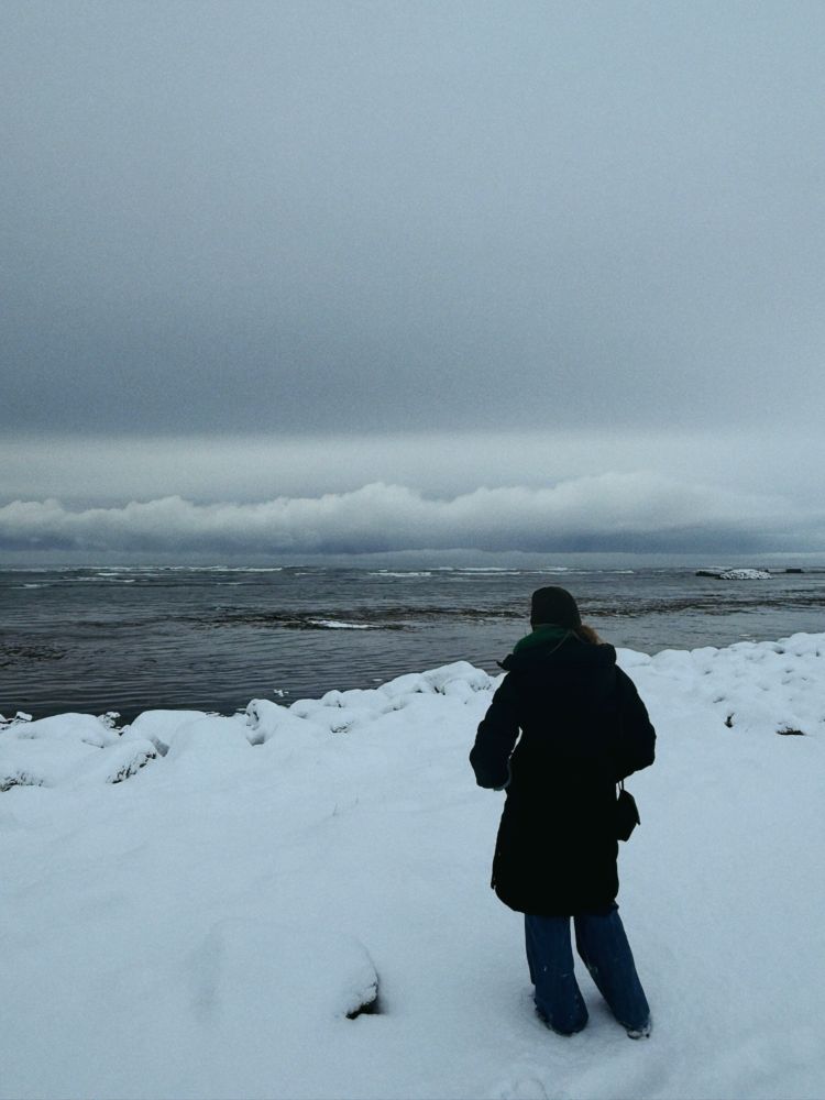 Person stands looking towards the sea, the beach covered in snow 
