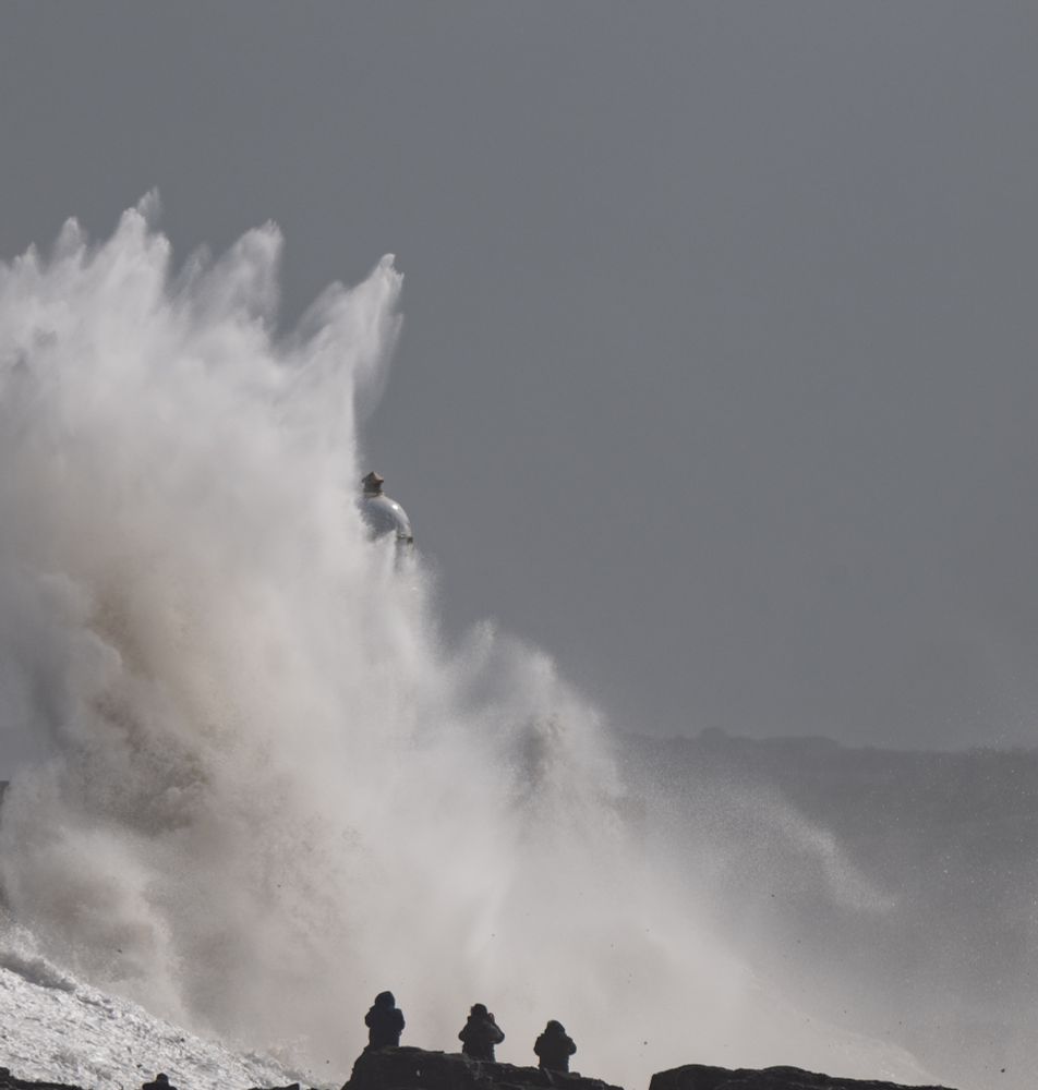 A wave bursts against the pier, dwarfing the 10m lighthouse on the end. There are 3 figures sat on rocks in the foreground - they look as though they are in danger but are actually quite some way from the sea