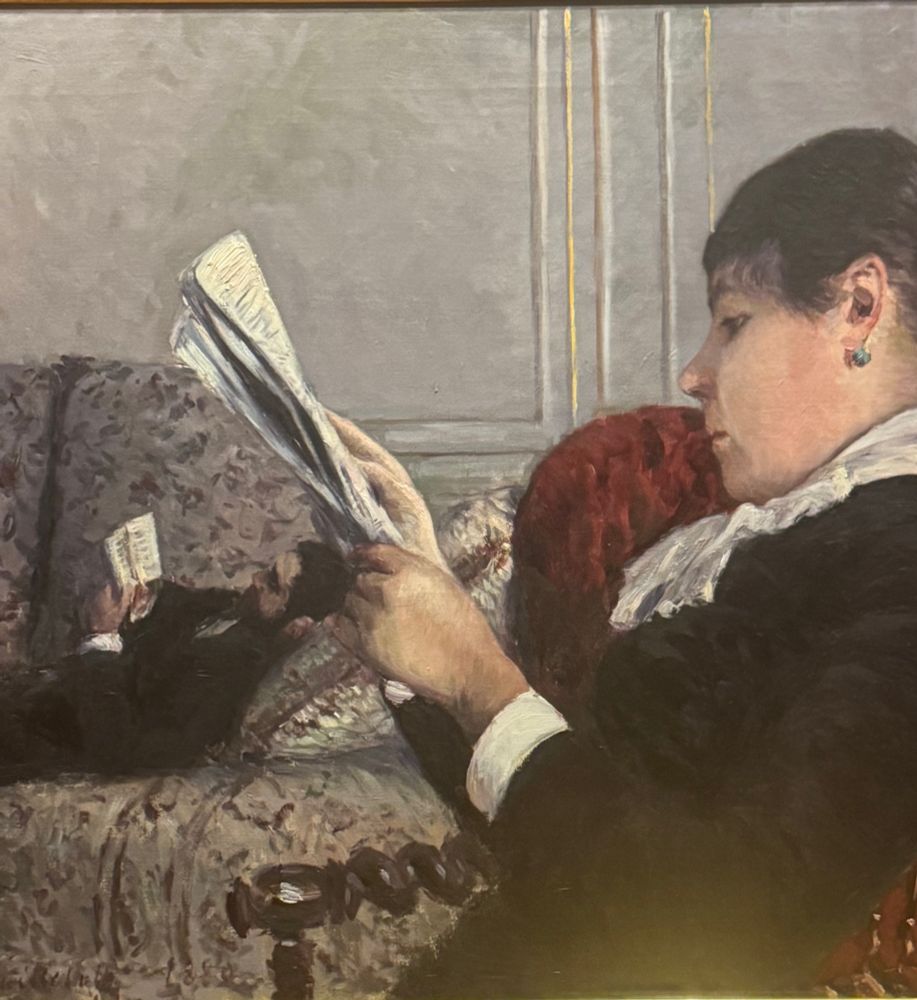 an impressionist painting with a gigantic woman reading a newspaper in the foreground, and in the background a much visibly smaller man reading a book on a couch. from the museum card:

Interior, Woman Reading
1880
Oil on canvas
A woman reads a newspaper in an armchair while a man reclines on a sofa with a novel. As he did in Interior, Woman at the Window (on view nearby), Caillebotte staged his bachelor friend Richard Gallo with an unidentified woman, perhaps Caillebotte's companion, Charlotte Berthier. This pictorial narrative both intrigued and irritated critics at the
1880 Impressionist exhibition: The dramatic shift in scale, with the woman dwarfing the man, was seen as emasculat-ing. Caillebotte further challenged gender stereotypes by showing the woman scouring a newspaper-then viewed as a masculine endeavor-while the lounging male reads a novel, likewise considered a feminine pastime.