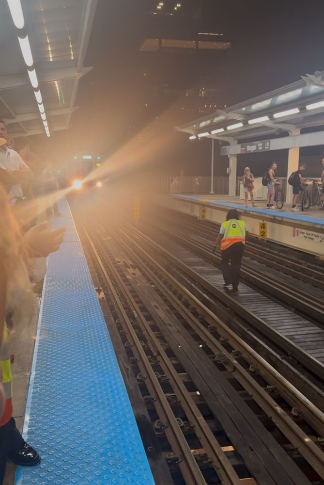 photo of the green line L platform. a CTA employee is down on the tracks, having retrieved a dropped phone. people on the edge of the frame can be seen clapping. an oncoming green train has stopped about 30 yards away while the employee gets off the tracks  