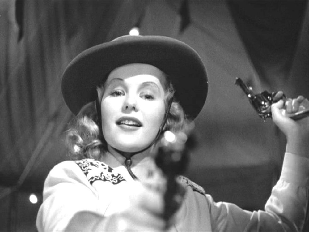 Peggy Cummins as a sideshow sharpshooter in Gun Crazy. With a gun cocked in her left hand, she smiled lazily with the gun in her right pointed directly at the viewer.