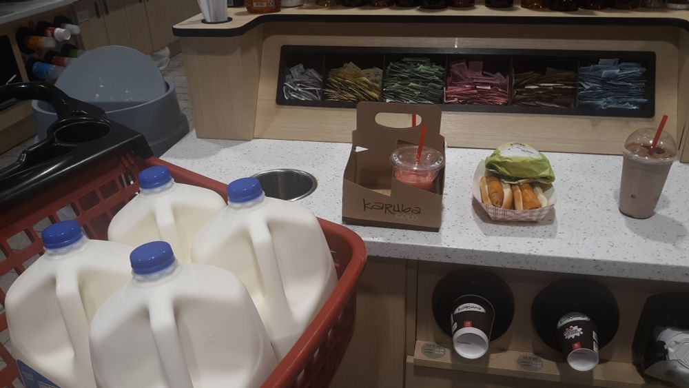 A photograph taken at the local 6th Street Kwik Stat gas station, taken in Marshalltown, Iowa, my hometown. Here, we have four milk jugs in a red grocery cart, followed by a mango F'real smoothie in the back (not seen because of the angle this was taken), a strawberry Fresh Blends' smoothie that are both sitting in a cupholder. To the right is a soon-to-be-loaded banana split chocolate Fresh Blends smoothie that was loaded with whipped cream, chocolate and cinnamon powder, as usual. The top part of the counter is cubbies for assorted items of sugar, sweetener, salt, etc., while in the far top left is a trash can that's partially half open. At the center portion of the counter is a quater pounder sandwich and two chicken and pepper Jack dogs in a hot dog holder I bought for generally myself. I was broke on paying for my snacks after this, since I now only have change leftover.