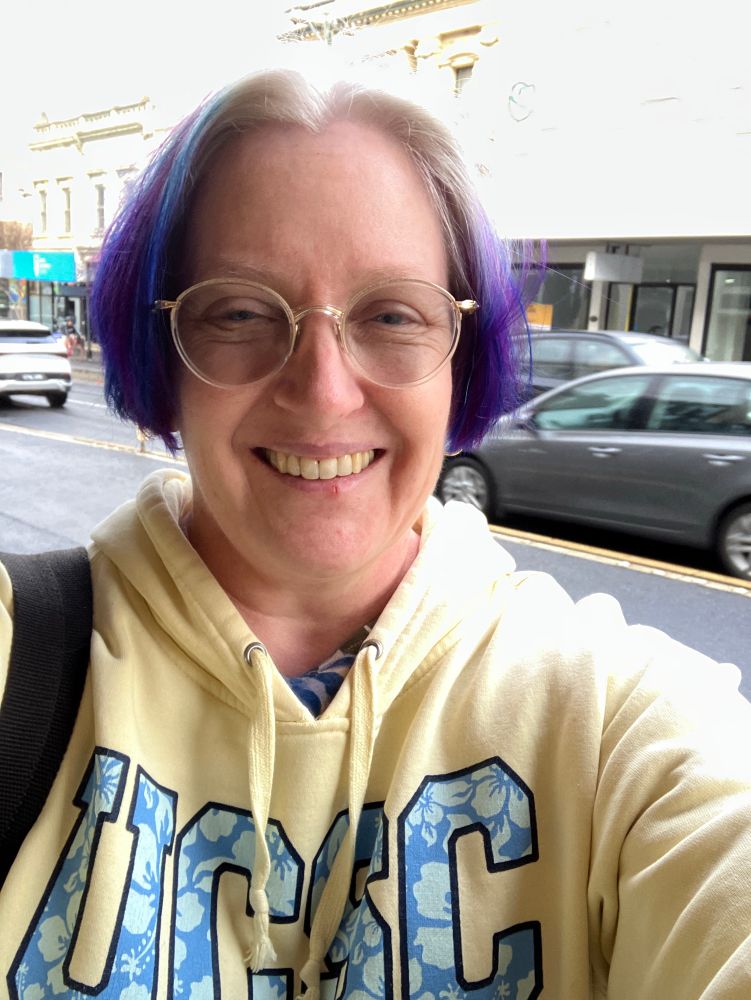 Selfie of me after emerging from the salon!   My hair is bobbed to my cheek level, white-blond at the roots with a mixed blue and purple ombre coming up from the tips.  I’m also wearing my glasses and my light yellow UCSC hoodie…hood down, of course!