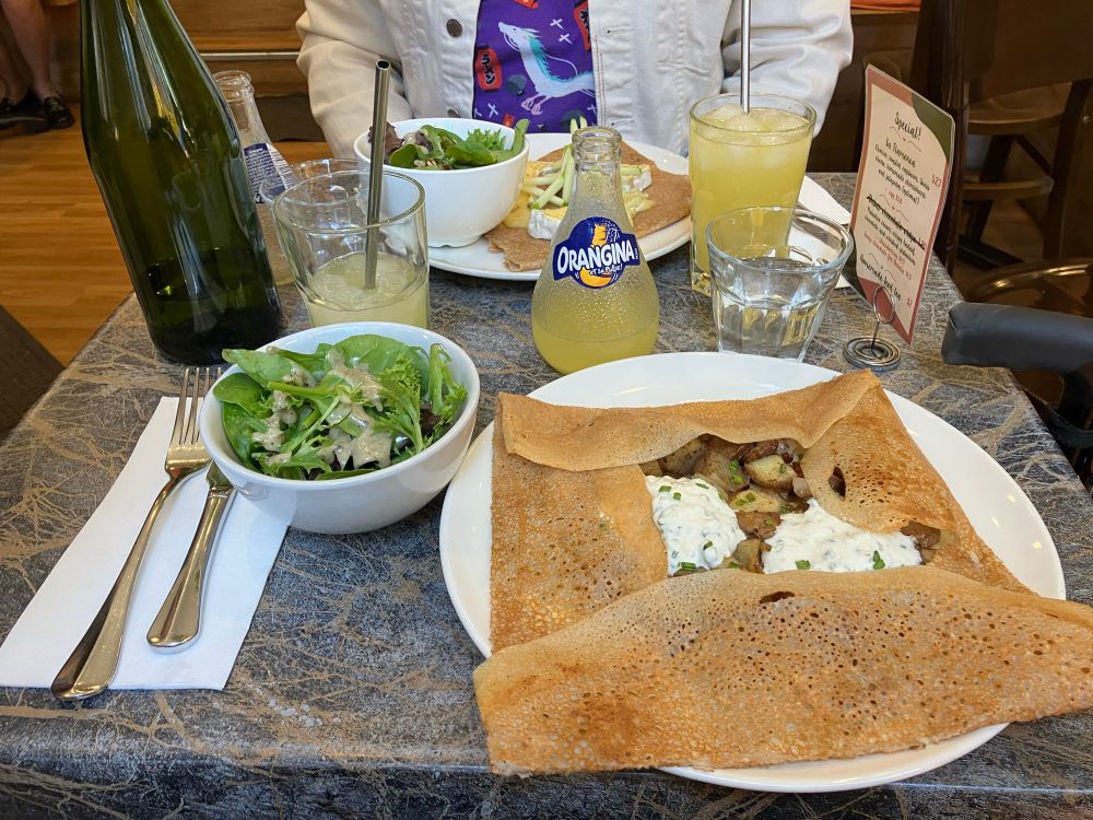 A cafe table set for two, focusing on a galette with spicy lamb and sausage and yogurt, and a green salad with mustard dressing in a small bowl. Also a bottle of Orangina!