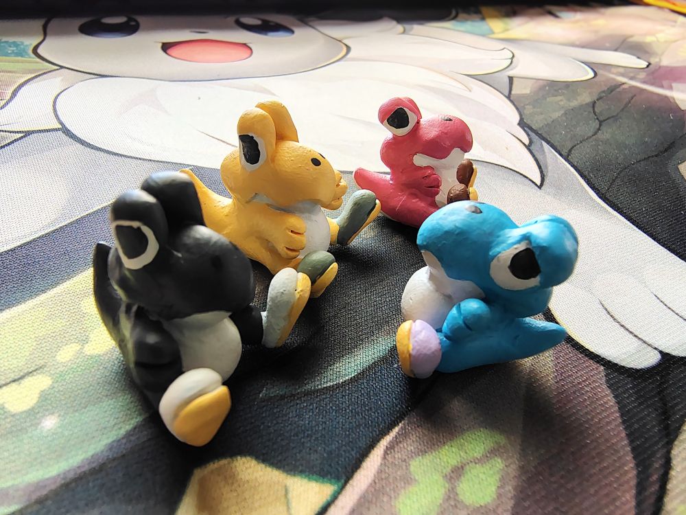 Four tiny clay baby yoshis. From left to right, they're black, yellow, pink, and light blue.