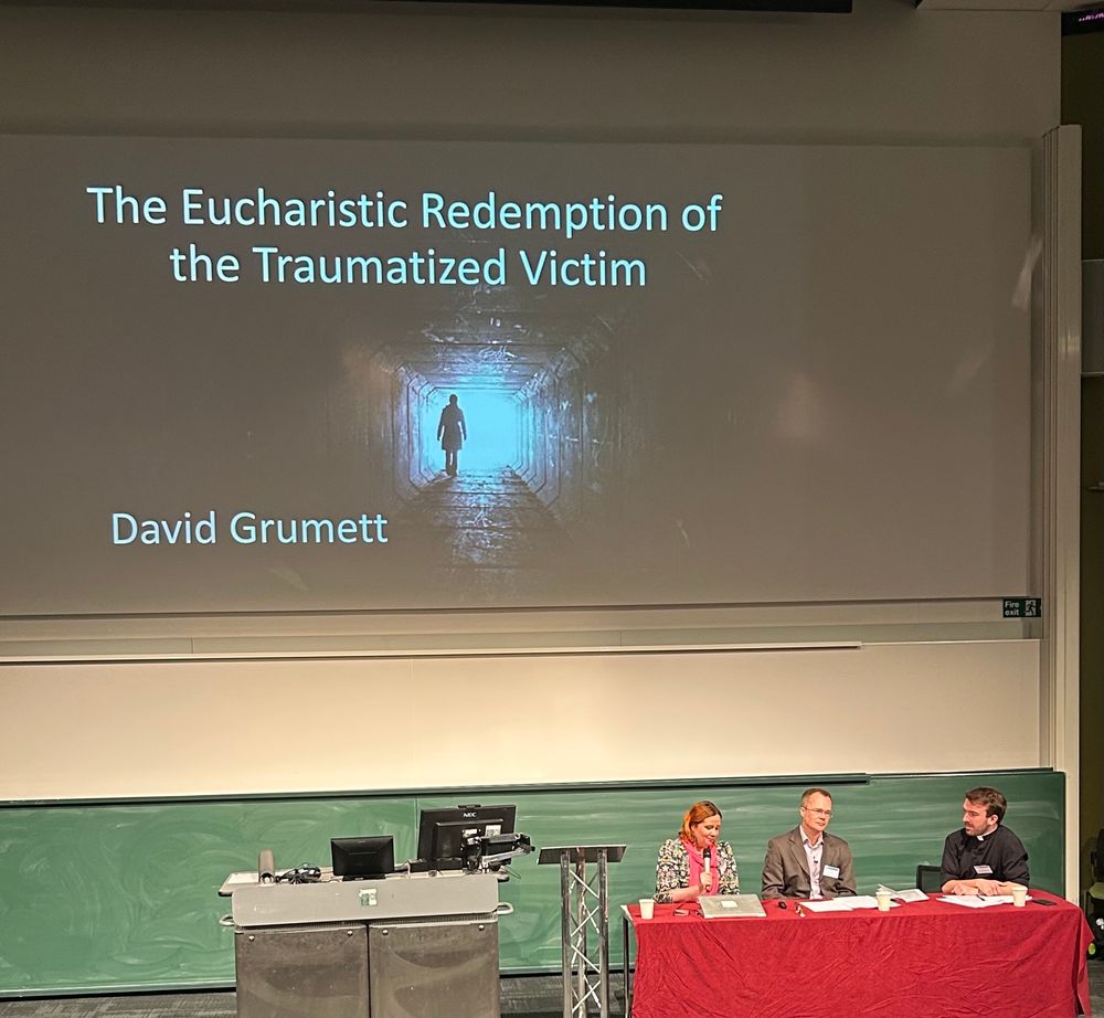 A panel of speakers (one woman and two men) sitting behind a red table. On the screen behind is the title of the talk “The Eucharistic Redemption of the Traumatized Victim” by David Grumett