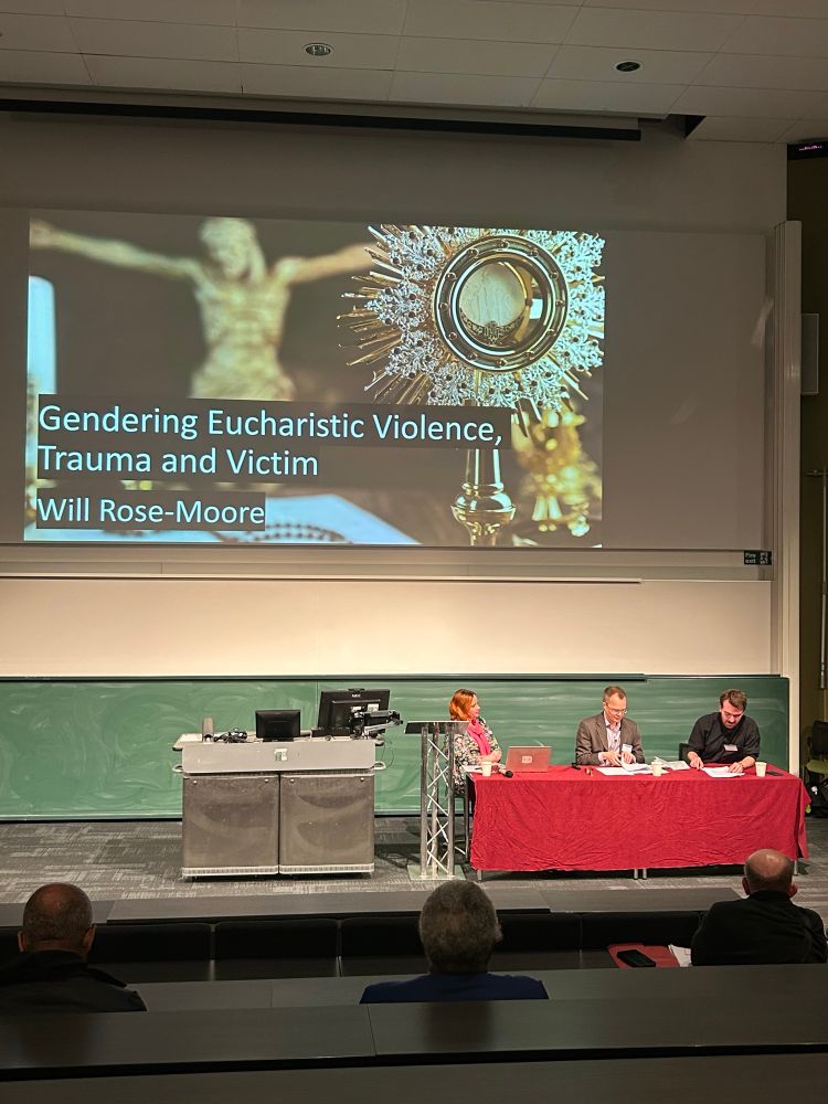 A panel of speakers (one woman and 2 men) sitting behind a red table. On the screen behind is the title of the talk “Gendering Eucharistic Violence, Trauma and Victim” by Will Rose-Moore