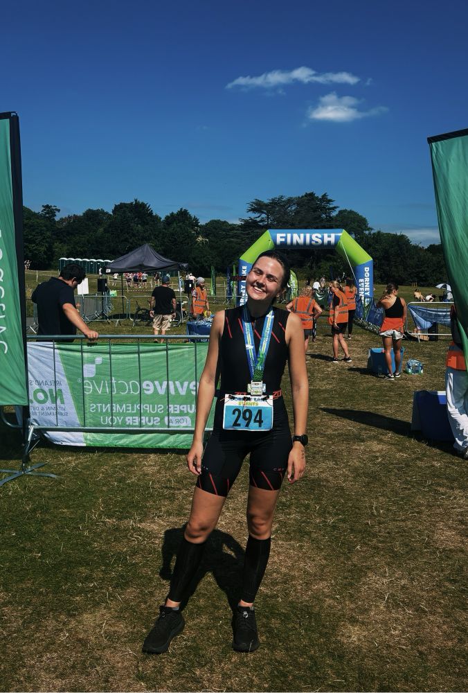 Moa is smiling and standing in front of a sign that says “Finish”. She is wearing her triathlon race number and triathlon suit.