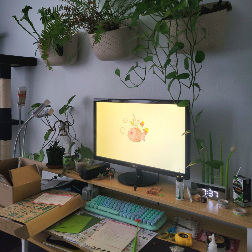 A small desk with plants, art and crafts supplies, stationery, packet of tomato seeds, soy milk and needle felted guinea pigs. Above the desk are a few wall planters with pothos, boston ivy and bird nest fern. Next to the desk is a black cat tree.
