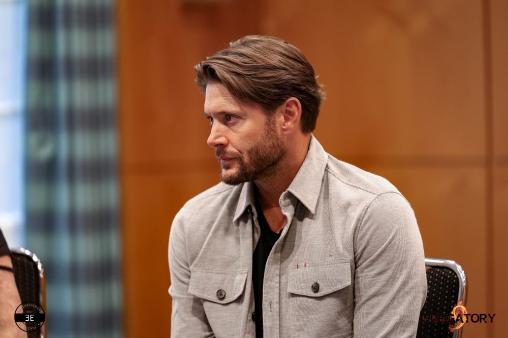 Jensen is sitting on a chair visible chest up. He is facing to the left of the camera and is listening with a focus on someone who is a bit more to the left. He is sitting up and leaning a bit forward. Hands are most likely on his lap.

He is wearing black jeans, black shirt and a light beige button up jacket with chest pockets. His sleeves are rolled up. Most of the background is a rustic colored wall, left side of the photo shows a stripped curtain and a bit of a big window