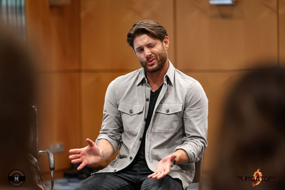 Jensen is sitting on a chair visible thighs up. He is facing to the left of the camera. He is mid explaining something while looking at someone to the right of the camera. He is making a face with his left eye closed and left side of mouth pulled up. His head is tilted to the right. His arms are in a 90 degrees bent in front of him, palms up. Both of his legs are on the ground

He is wearing black jeans, black shirt and a light beige button up jacket with chest pockets. His sleeves are rolled up. Background wall is rustic in color.